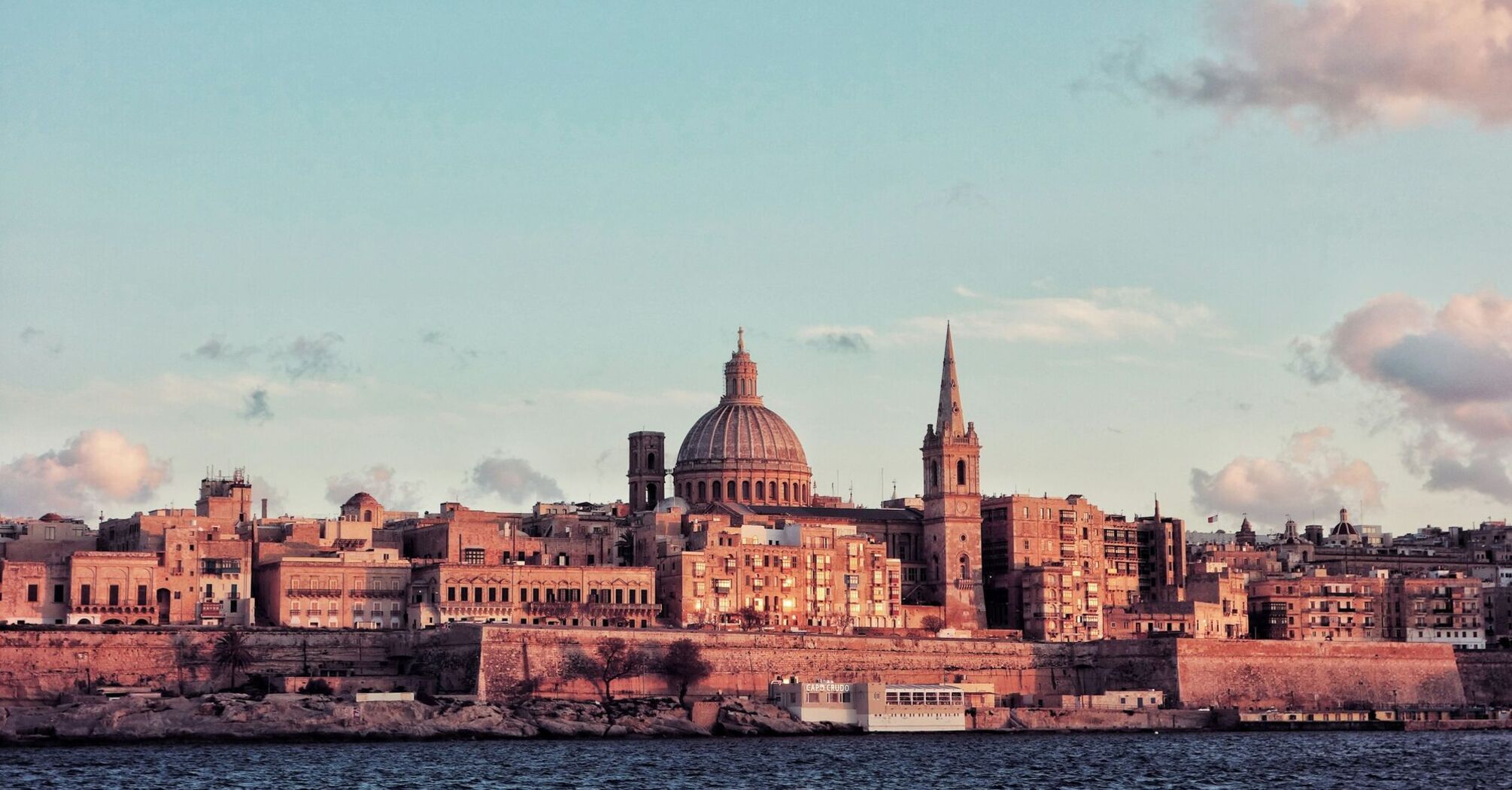 A scenic view of Valletta, Malta, featuring its historic domed cathedral and spired church bathed in soft sunset light