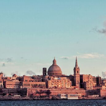 A scenic view of Valletta, Malta, featuring its historic domed cathedral and spired church bathed in soft sunset light