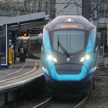 A TransPennine Express train at a station platform with passengers nearby, displaying "Manchester Airport" on its destination board