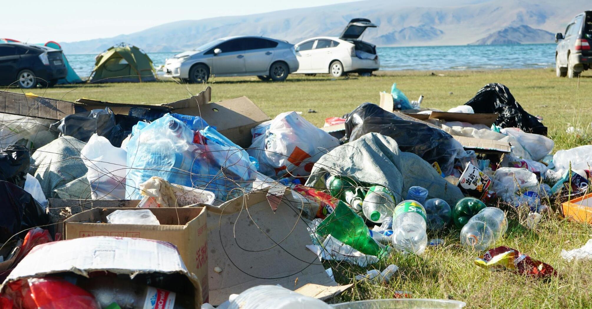 Trash scattered on grass near a lakeside camping area with cars and tents in the background