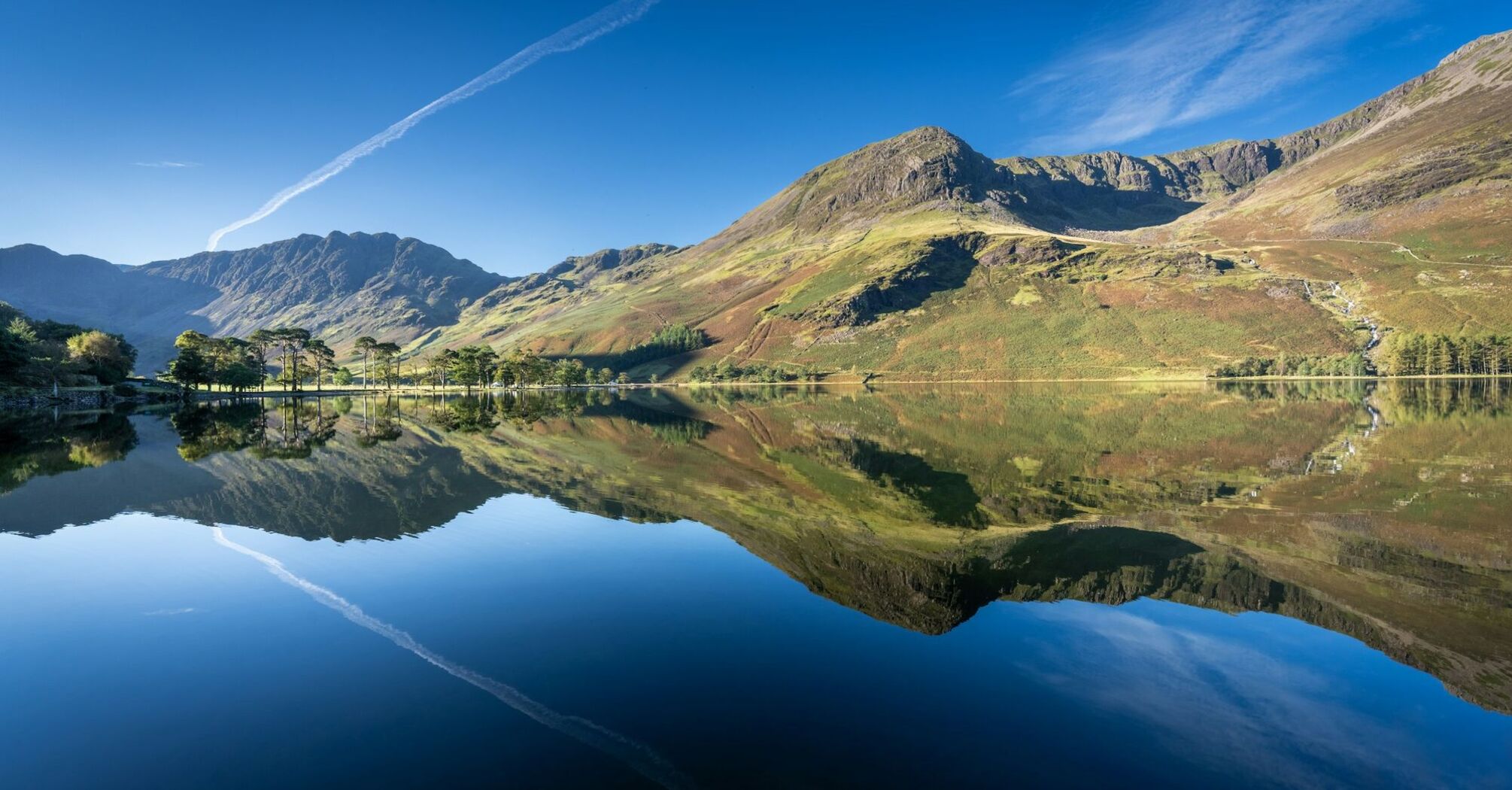 A calm lake in the Lake District reflects surrounding mountains under a clear blue sky