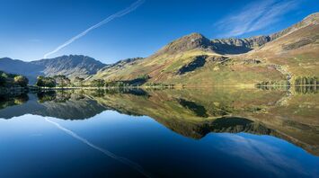 A calm lake in the Lake District reflects surrounding mountains under a clear blue sky