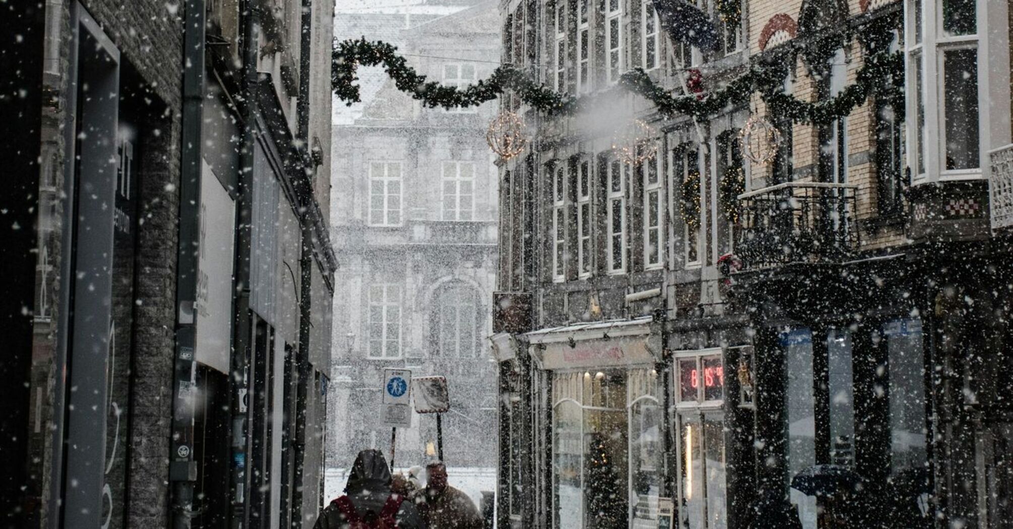 A snowy Munich street with historic buildings decorated for the winter season, featuring people walking under garland-adorned streets