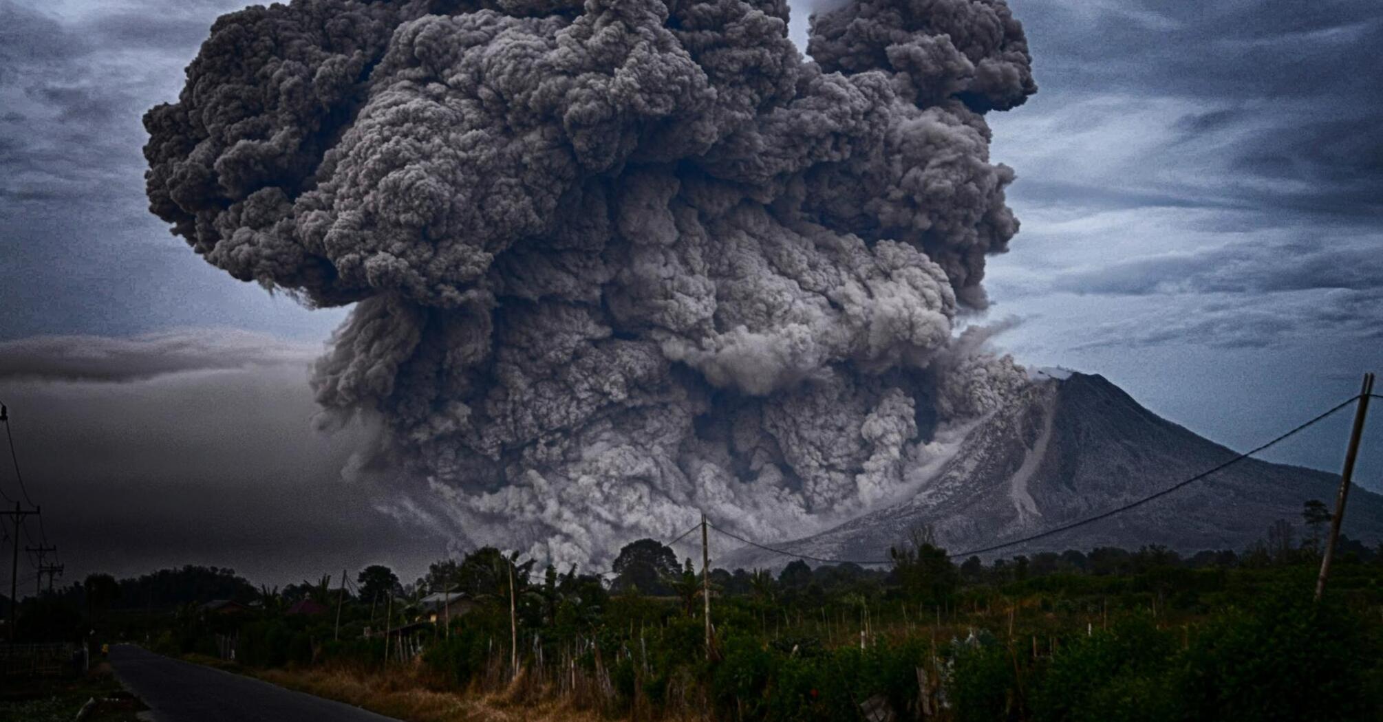 A massive eruption with thick dark ash clouds billowing from a volcanic mountain under a stormy sky