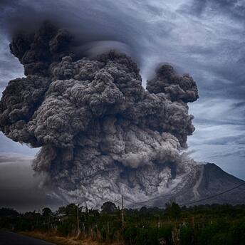 A massive eruption with thick dark ash clouds billowing from a volcanic mountain under a stormy sky