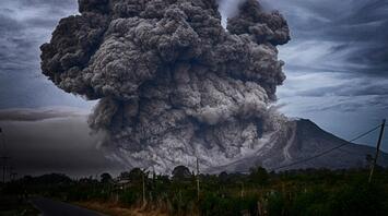 A massive eruption with thick dark ash clouds billowing from a volcanic mountain under a stormy sky