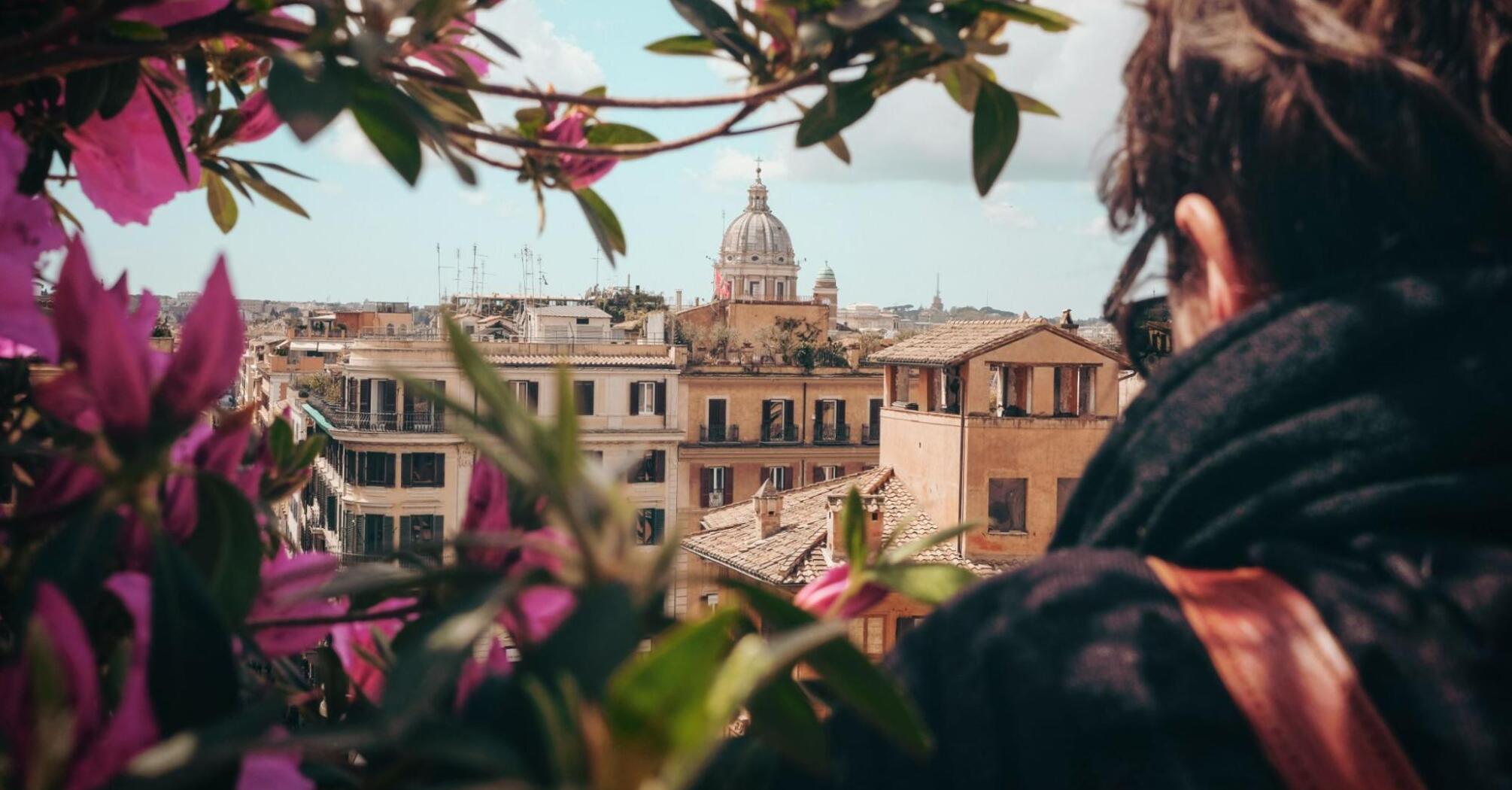 View of historic buildings and domed church in Italy, framed by pink flowers, with a person observing the scene