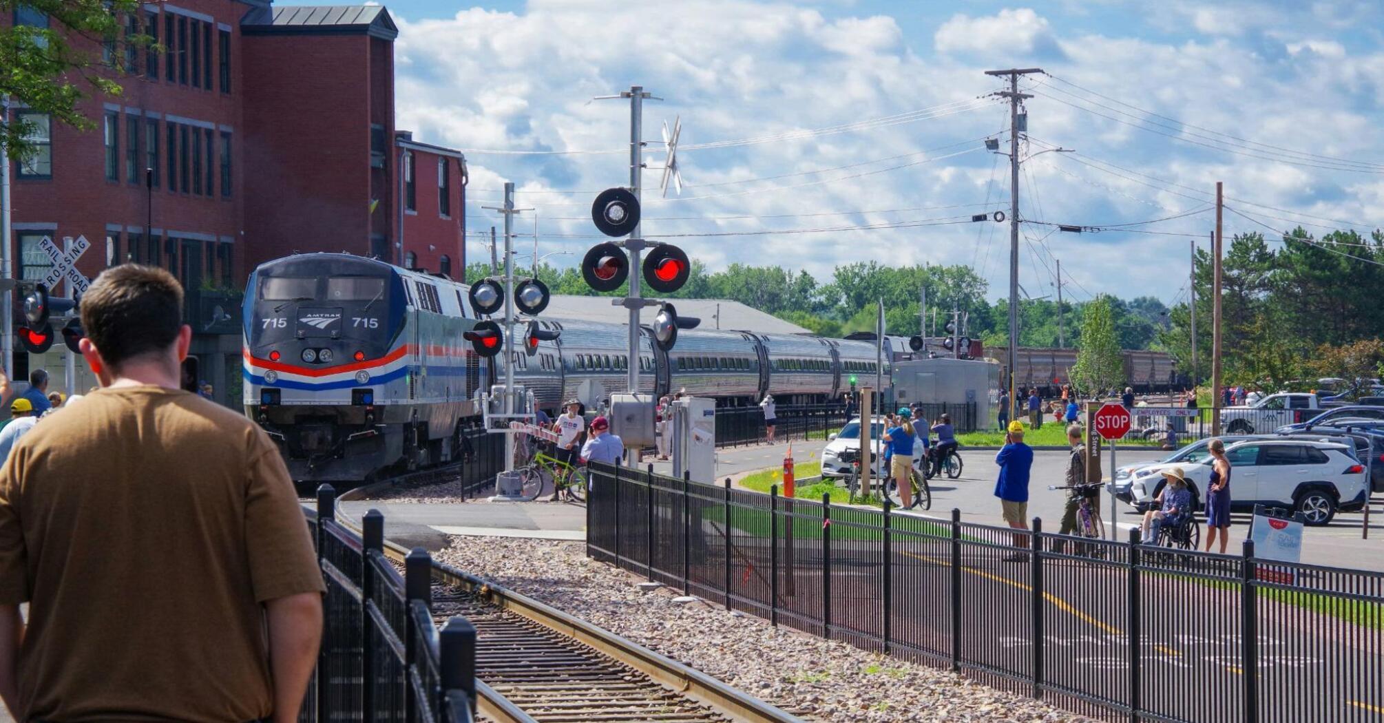 Amtrak train crossing a busy intersection as people watch and wait nearby on a clear, sunny day