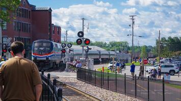 Amtrak train crossing a busy intersection as people watch and wait nearby on a clear, sunny day