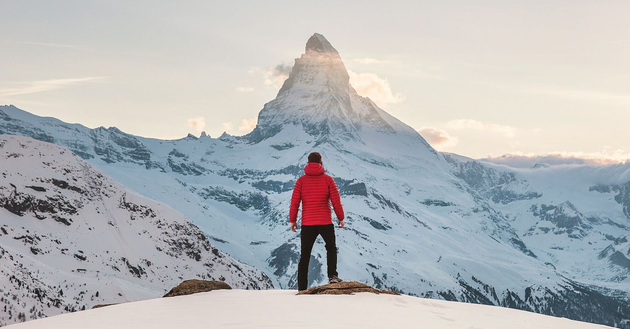 A traveler in a red jacket stands facing mountain in a snowy winter landscape