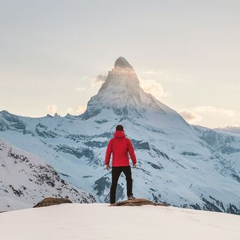 A traveler in a red jacket stands facing mountain in a snowy winter landscape