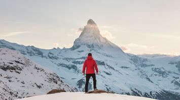 A traveler in a red jacket stands facing mountain in a snowy winter landscape