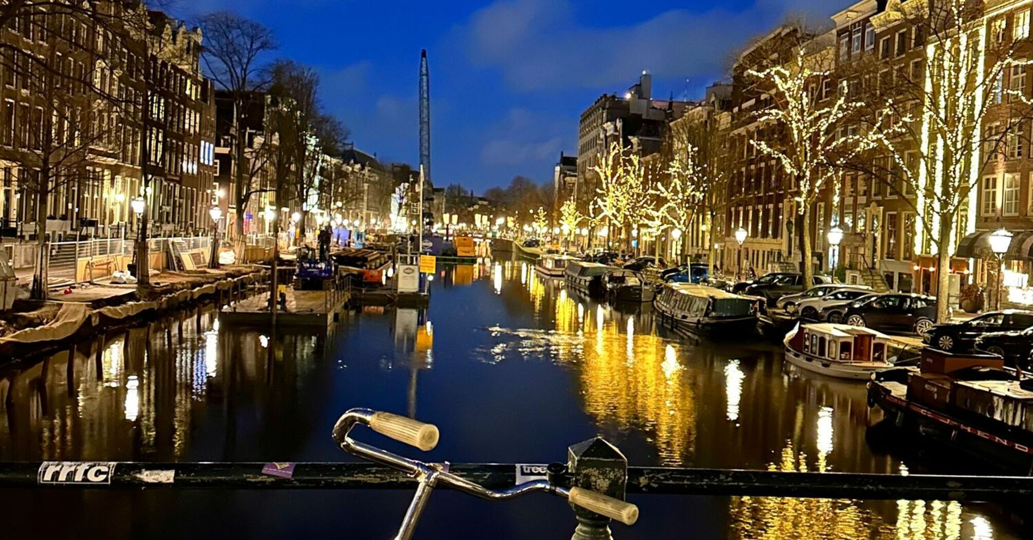 A bicycle parked on a bridge overlooking a lit Amsterdam canal at night, with illuminated trees and buildings reflecting in the water