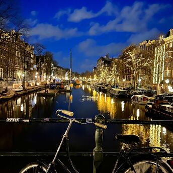 A bicycle parked on a bridge overlooking a lit Amsterdam canal at night, with illuminated trees and buildings reflecting in the water