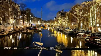 A bicycle parked on a bridge overlooking a lit Amsterdam canal at night, with illuminated trees and buildings reflecting in the water