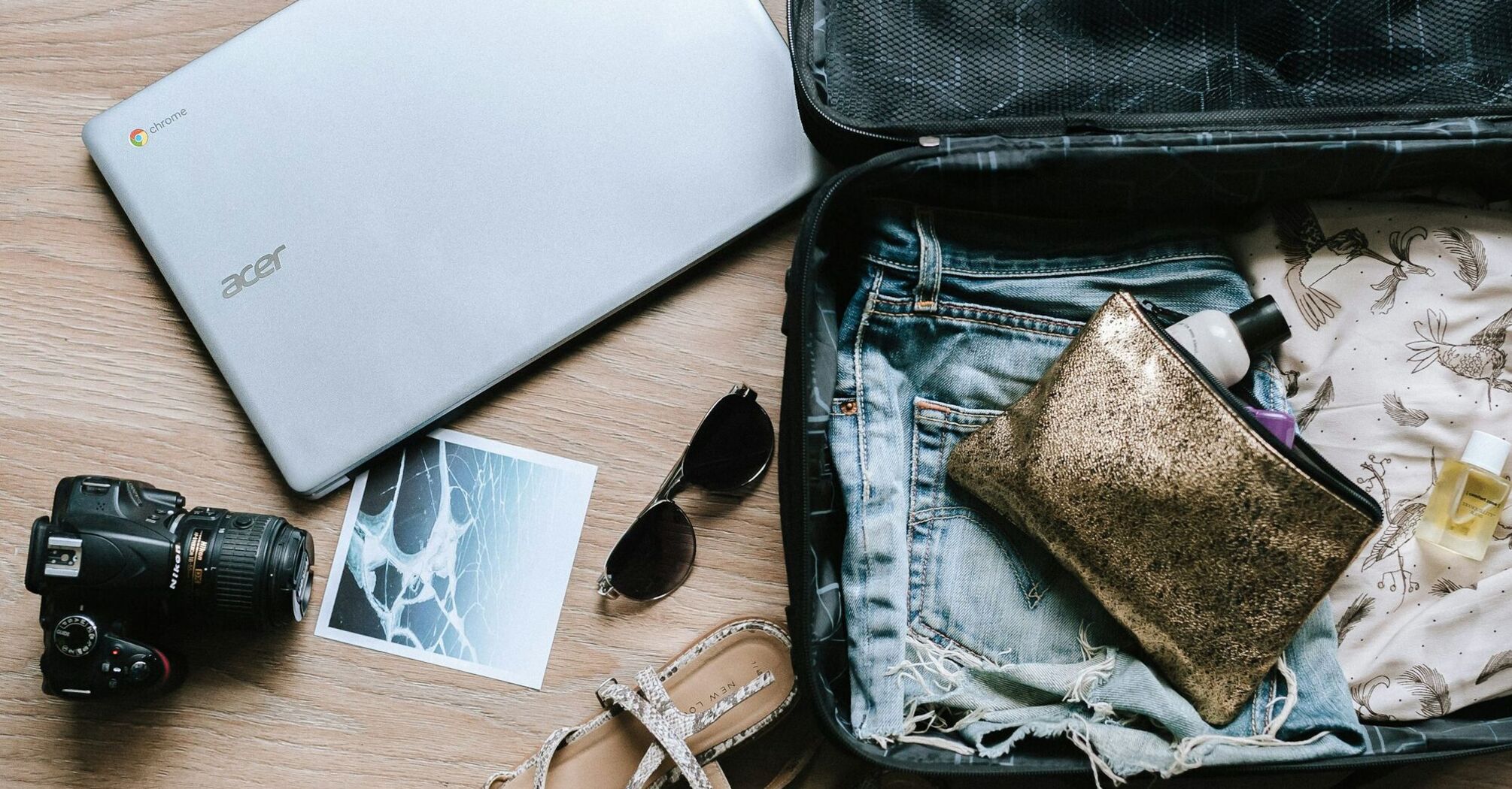 An open suitcase with travel essentials, including denim shorts, a camera, sunglasses, and a laptop, placed on a wooden floor