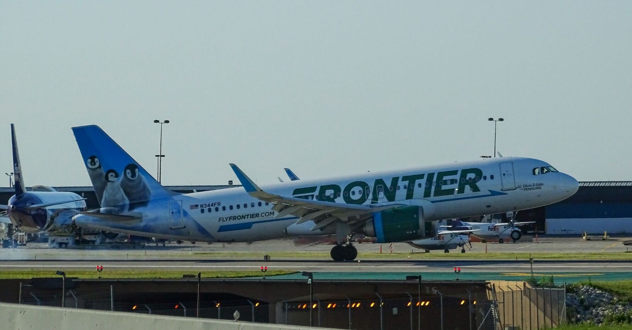 Frontier Airlines aircraft parked at an airport runway with penguin artwork on its tail