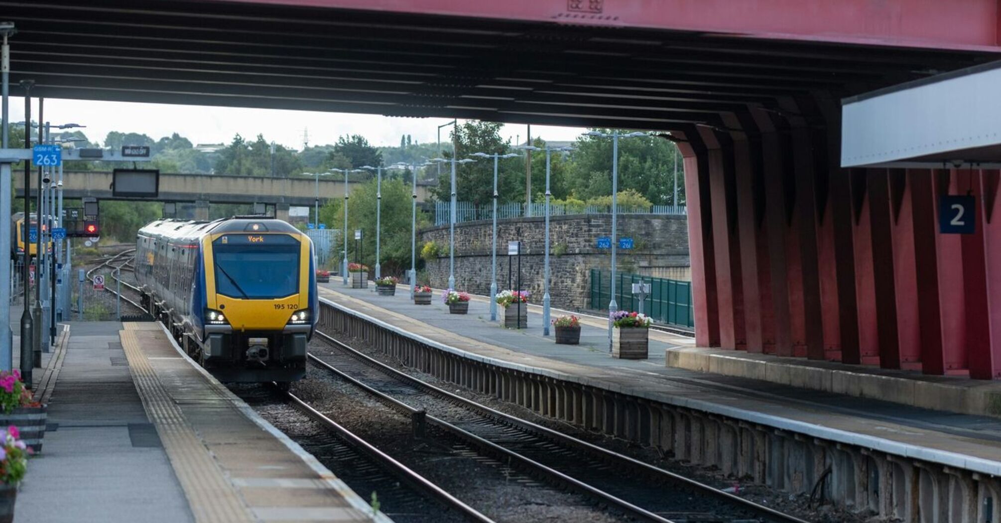 A train arrives at a platform under a large red bridge at a quiet station