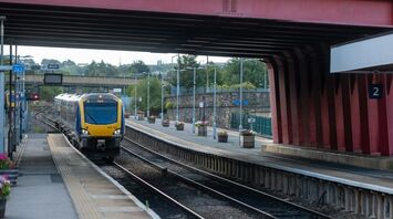 A train arrives at a platform under a large red bridge at a quiet station