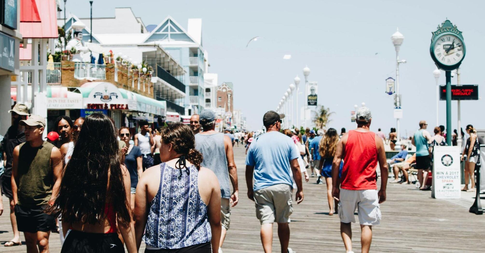 A crowded boardwalk filled with people enjoying a sunny day, with shops and signs in the background