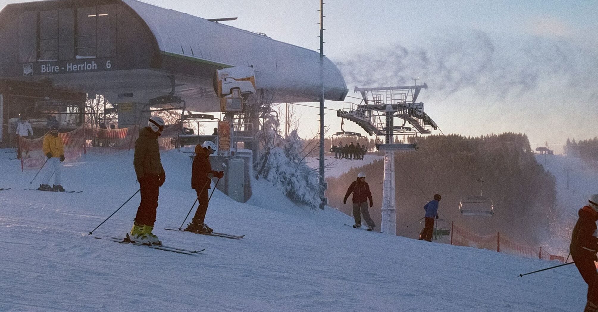 Skiers prepare at the base of a snowy slope near a ski lift station during sunrise