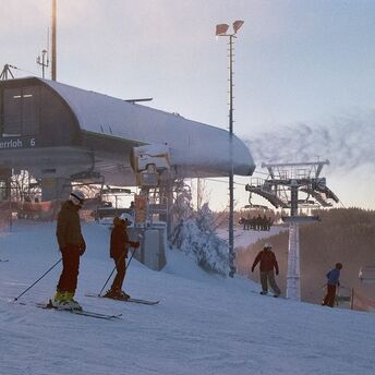 Skiers prepare at the base of a snowy slope near a ski lift station during sunrise