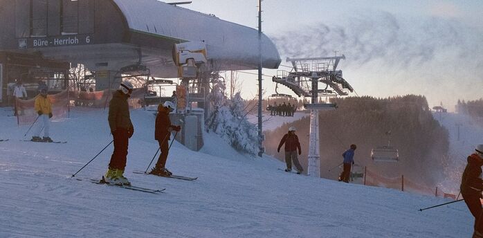 Skiers prepare at the base of a snowy slope near a ski lift station during sunrise