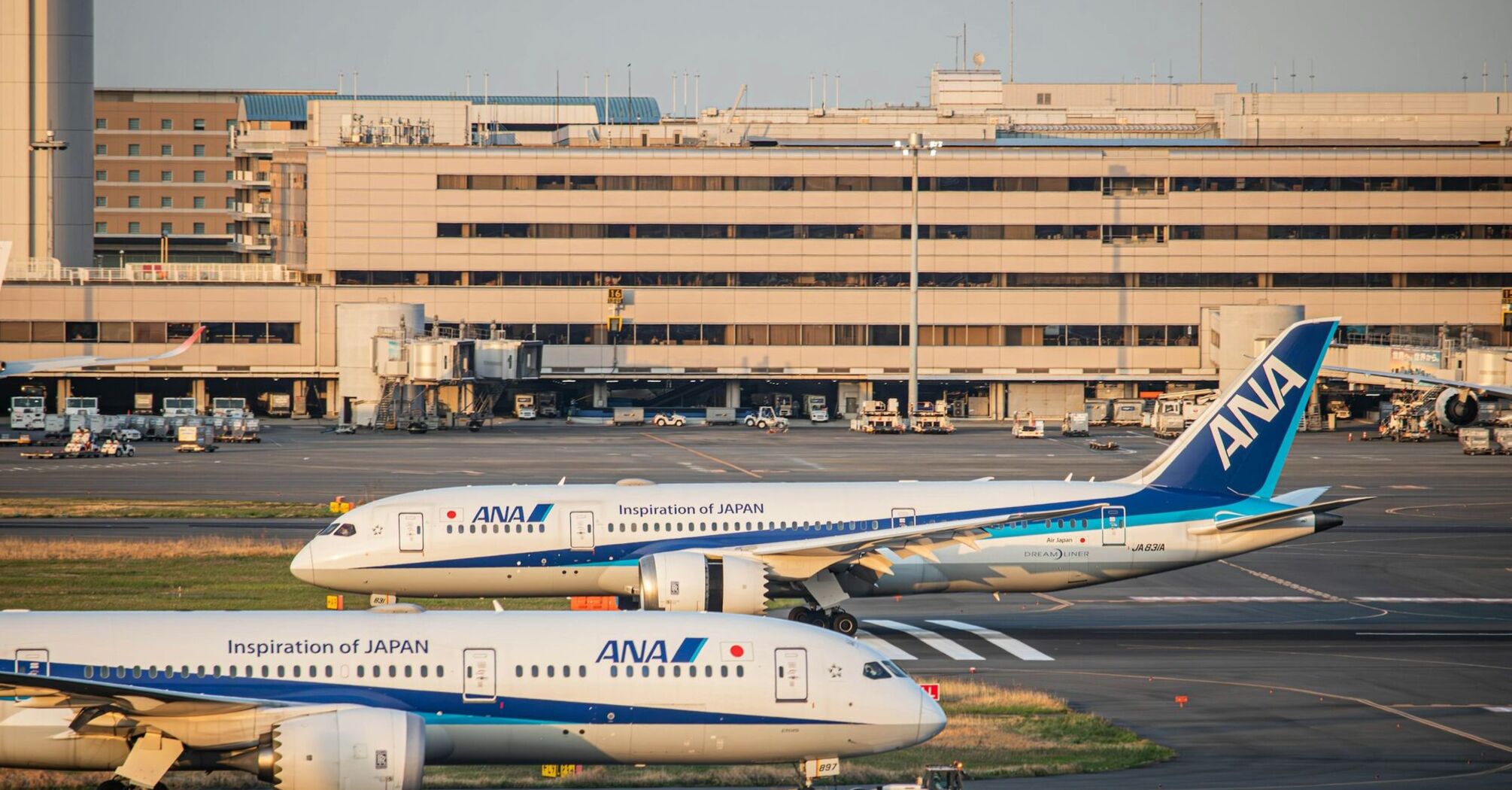 Two ANA aircraft parked at Haneda Airport, Tokyo, with terminal buildings and boarding gates in the background during sunset