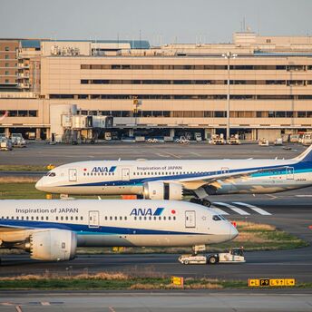 Two ANA aircraft parked at Haneda Airport, Tokyo, with terminal buildings and boarding gates in the background during sunset