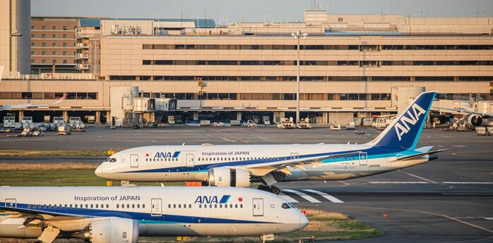 Two ANA aircraft parked at Haneda Airport, Tokyo, with terminal buildings and boarding gates in the background during sunset