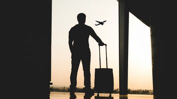 Silhouette of a traveler with luggage at an airport window, airplane taking off in the background