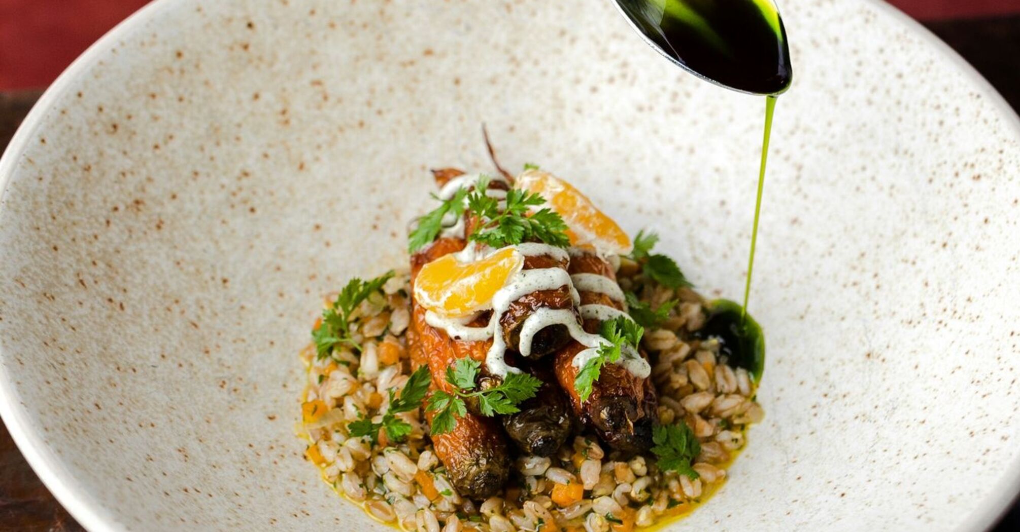 A hand drizzling green oil over a gourmet dish featuring roasted carrots, grains, and herbs in a rustic ceramic bowl