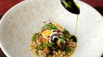 A hand drizzling green oil over a gourmet dish featuring roasted carrots, grains, and herbs in a rustic ceramic bowl