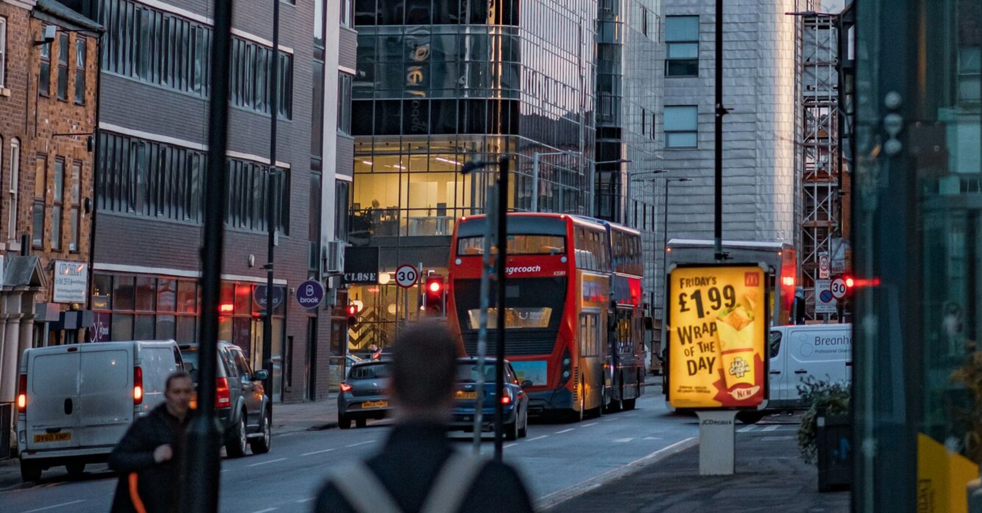 A busy city street in Greater Manchester with modern buildings, a red double-decker bus, and illuminated advertisements at dusk