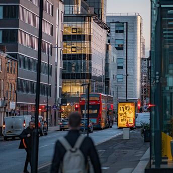 A busy city street in Greater Manchester with modern buildings, a red double-decker bus, and illuminated advertisements at dusk
