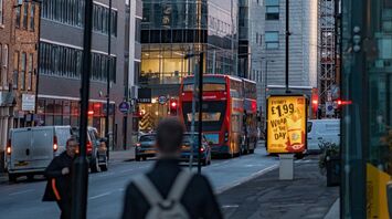 A busy city street in Greater Manchester with modern buildings, a red double-decker bus, and illuminated advertisements at dusk