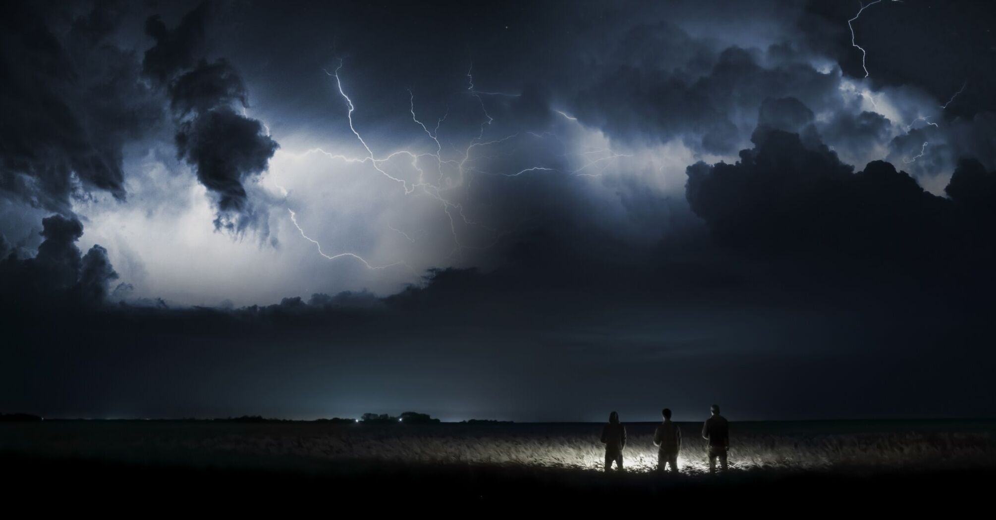 A stormy night sky with lightning illuminating clouds and silhouettes of three people standing in a field