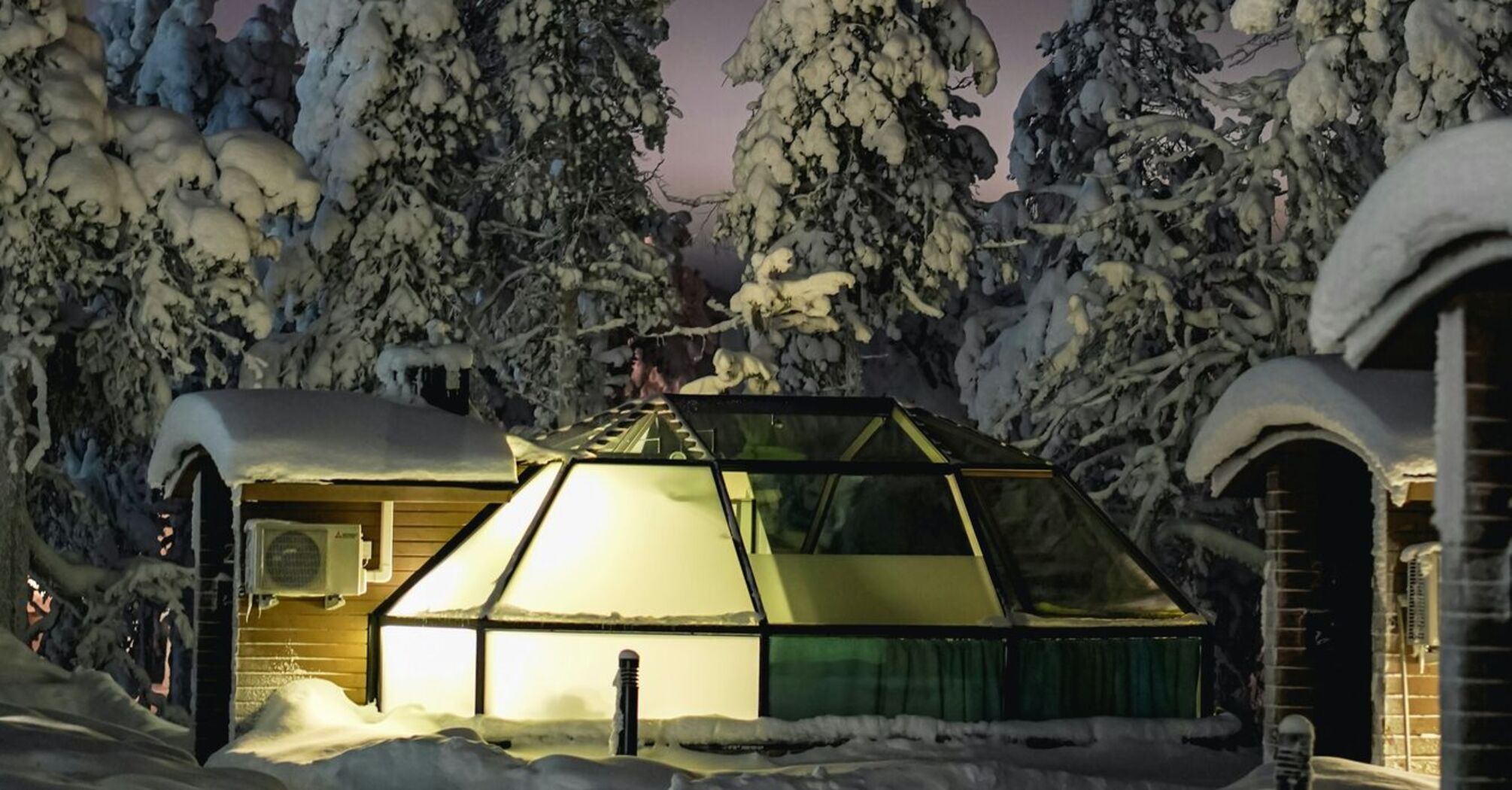 A glass igloo illuminated at night amidst snow-covered trees in Lapland