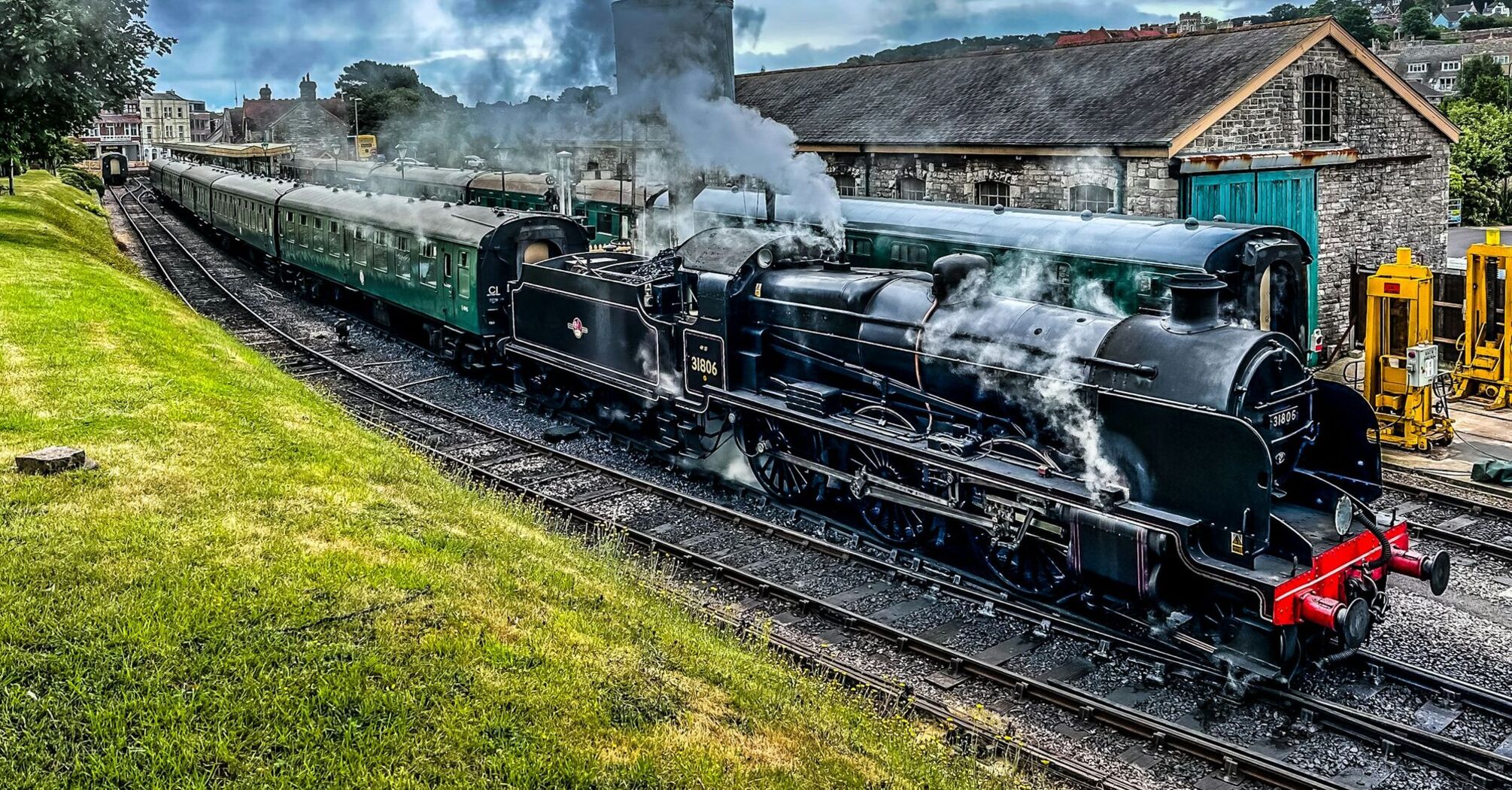 A historic Swanage Railway steam locomotive with passenger carriages near a vintage railway building