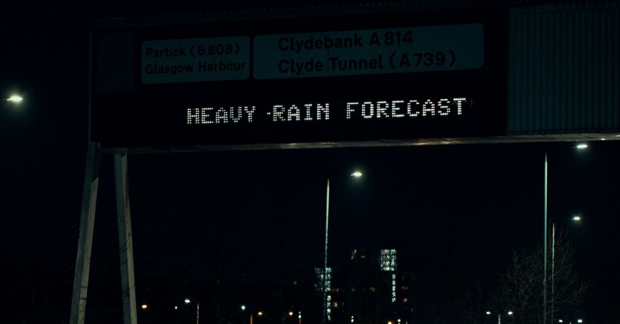 A digital road sign at night warning of heavy rain, with wet roads and passing cars visible