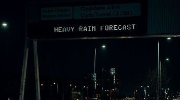 A digital road sign at night warning of heavy rain, with wet roads and passing cars visible