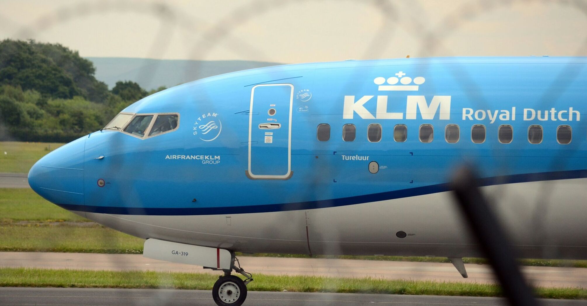 A KLM Royal Dutch Airlines plane taxiing on a runway with a blurred barbed wire fence in the foreground