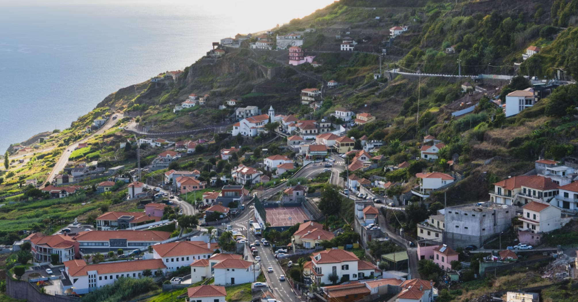 A scenic view of a coastal village in Madeira, showcasing terraced hills, red-roofed houses, and the ocean in the background