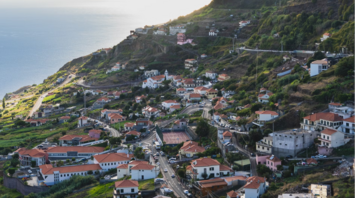 A scenic view of a coastal village in Madeira, showcasing terraced hills, red-roofed houses, and the ocean in the background