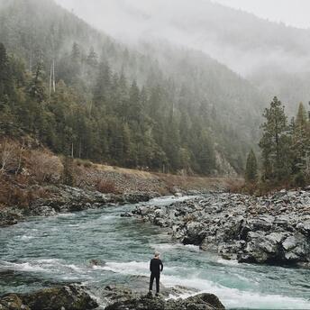 A person standing by a river in a misty forest landscape, surrounded by rocks and dense trees