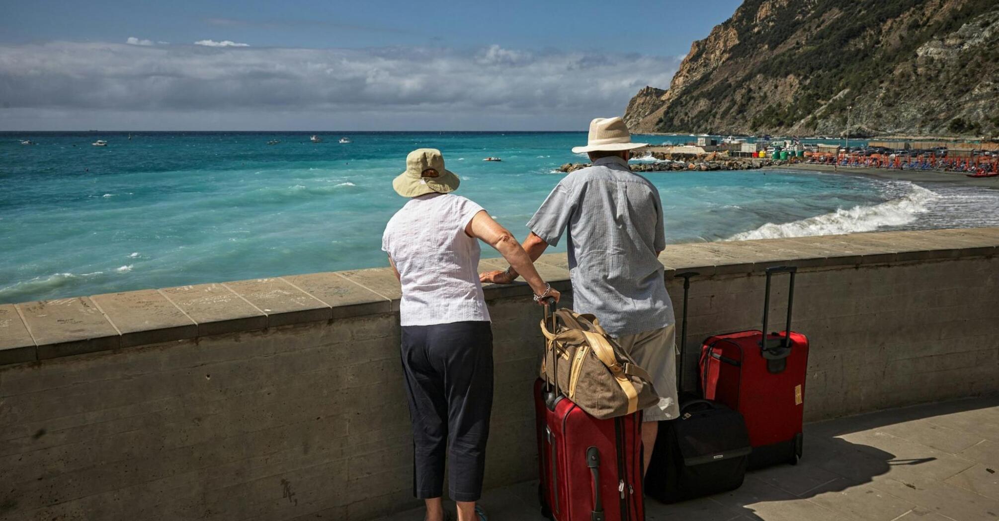 Two elderly travelers with luggage admire a seaside view, standing by a coastal promenade with a scenic backdrop