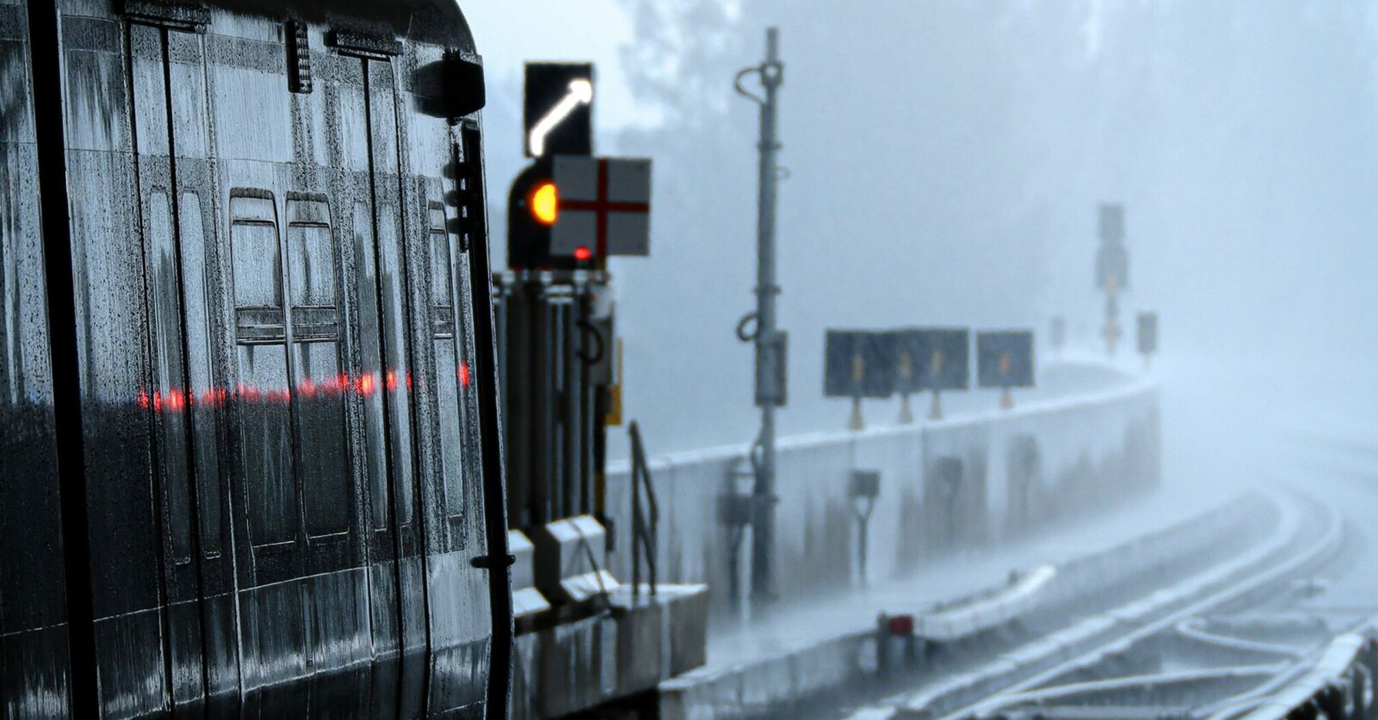 A train station platform during heavy rainfall, with wet tracks and misty surroundings