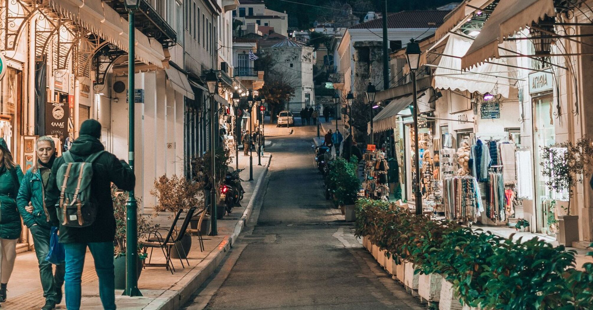 A picturesque street in Athens with a view of the Acropolis in the background, lined with local shops and evening lights