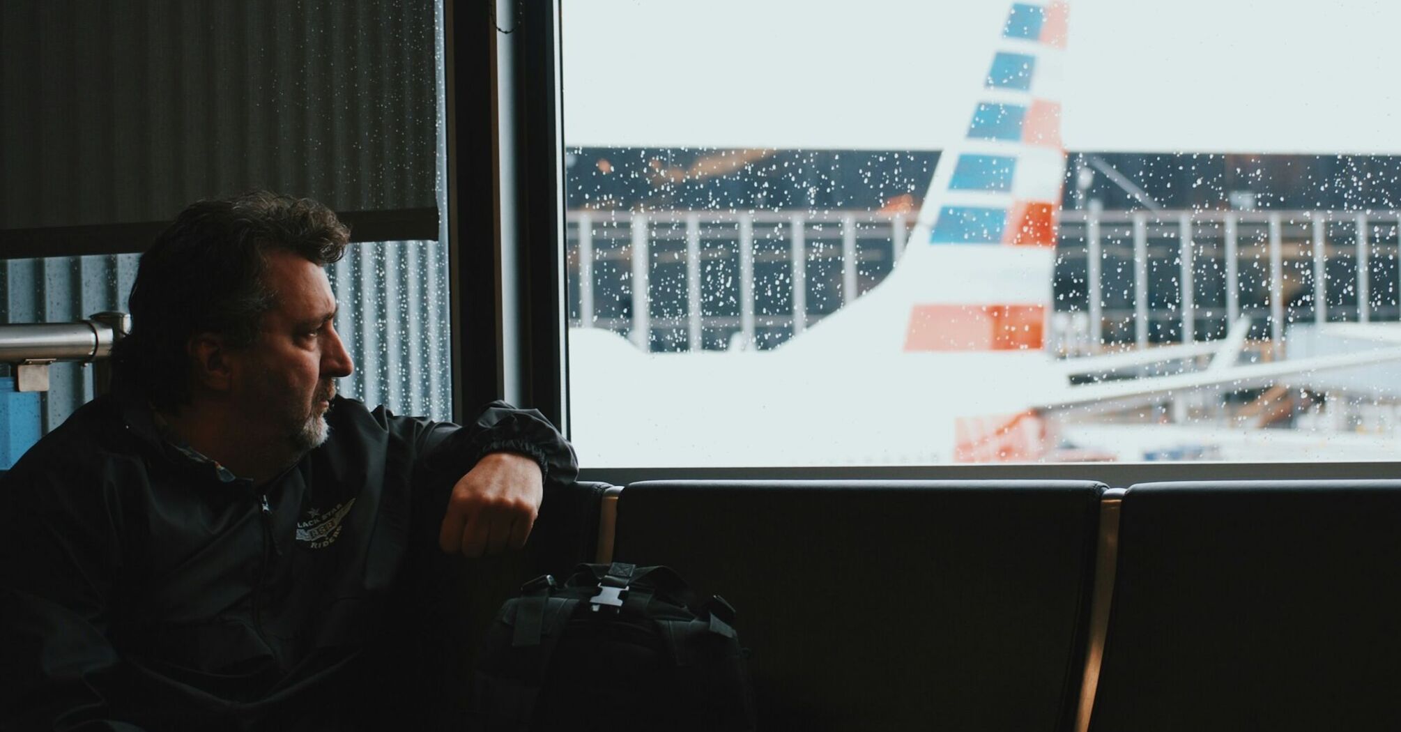 A man sitting by a window at an airport, looking out at a plane in rainy weather