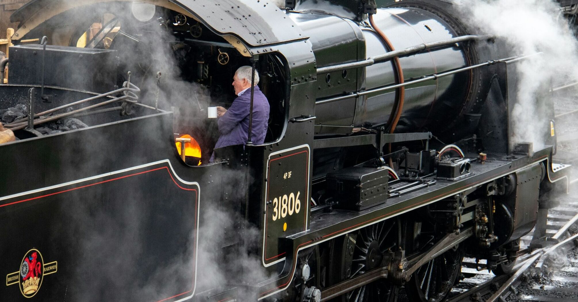 A heritage steam locomotive in operation with a fireman tending to the firebox, surrounded by steam